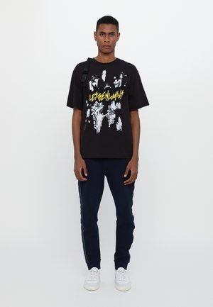 Schwarzes Grafik-T-Shirt mit weißem und gelbem Text sowie abstrakten Designs; kombiniert mit marineblauen Jogginghosen und weißen Sneakern. Schlichter, lässiger Stil.