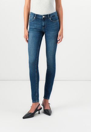 Kvinde iført tætsiddende blå jeans og sorte kitten heel slingback sko, stående mod en ensfarvet hvid baggrund.