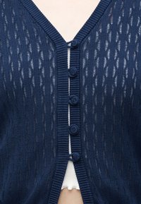 Cardigan en tricot bleu marine avec motif texturé, cinq boutons recouverts de tissu, porté sur un top côtelé blanc visible en dessous.
