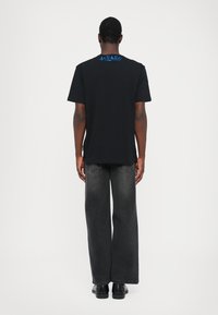 Svart bomullst-shirt med blå "MILANO"-text på ryggen, kort ärm, rak skärning, ihopparad med grå vida jeans och svarta skor.