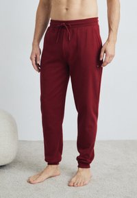 Mann in maroon Tommy Hilfiger Jogginghose, barfuß auf einem hellen Teppich stehend, mit einem hellen, strukturierten Stuhl in der Nähe.