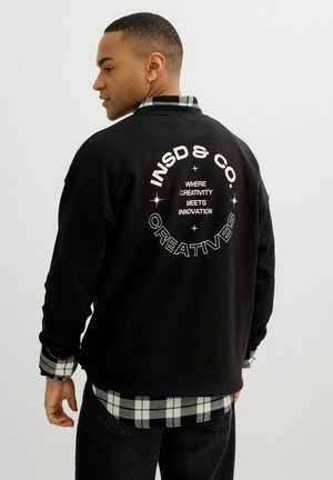 Hombre que lleva una sudadera negra sobre una camisa de cuadros en blanco y negro, con texto blanco "INSD & CO. CREATIVES" y "WHERE CREATIVITY MEETS INNOVATION" en la espalda.