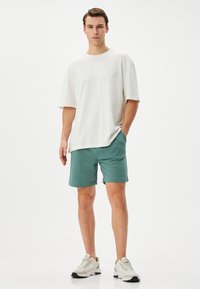Witte katoenen T-shirt met korte mouwen, gecombineerd met turquoise shorts en lichte sneakers met donkere zolen. Model staat met handen in de zakken.