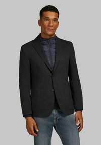 Zwarte op maat gemaakte blazer met twee voorzakken en een enkele knoopsluiting, gelaagd over een marineblauwe gewatteerde jack met rits, gedragen met blauwe spijkerbroeken.