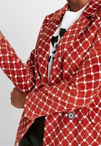 Röd kordflorensblazer med ett mönster av vita hjärtan och kedjor, snedskurna slag, två knappar och en lätt texturerad yta.