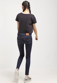 Jeans skinny azul oscuro con costuras contrastantes, cintura alta y una insignia de cuero en la parte trasera. Combinados con una camiseta negra y zapatillas plateadas.