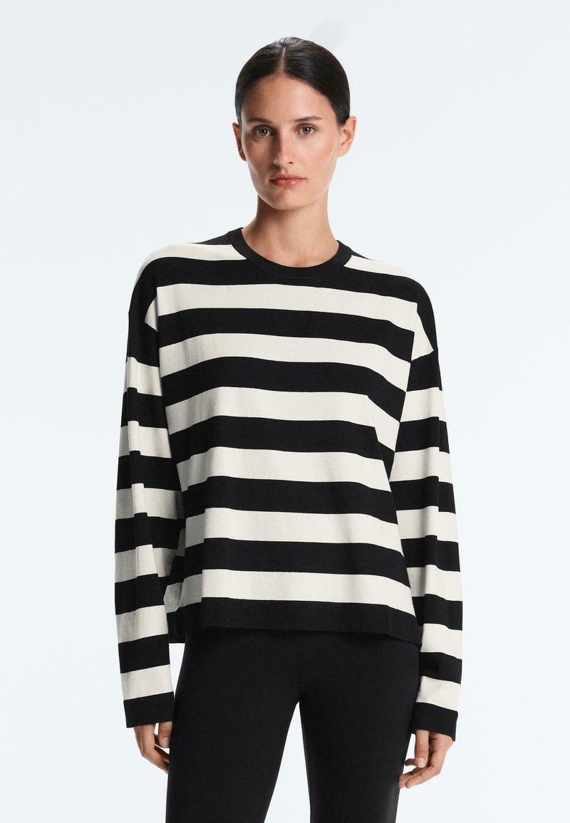 Femme aux cheveux foncés portant un haut à manches longues avec des rayures horizontales noires et blanches et un pantalon noir, debout devant un fond clair uni.