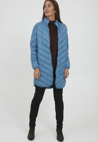 Ljusblå vadderad jacka med hög krage, diagonalt quiltat mönster, dragkedja fram och långa ärmar, kombinerad med svarta slim fit-byxor och bruna stövlar.