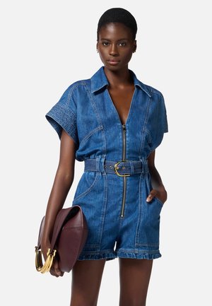 Tutina in denim con maniche corte, zip frontale e cintura con fibbia dorata. Il modello tiene una borsa bordeaux con dettagli dorati.