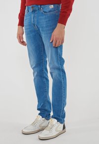 Uomo che indossa jeans slim-fit azzurri, sneakers bianche e beige e una maglietta rossa a maniche lunghe, in piedi contro uno sfondo neutro.
