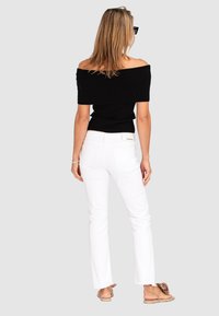 Black off-the-shoulder ribbed top paired with white flared pants. Model wears brown flat sandals with fabric floral accent. Neutral background.
