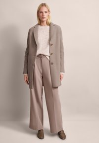 Beige Wollmischungsjacke mit einer Reverskragen und drei Knöpfen, kombiniert mit einem hellbeigen Pullover und weiten Hosen in einem passenden Farbton.