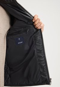 Una mano tiene aperta la parte interna di una giacca di pelle nera, mostrando una tasca con zip e l'etichetta "JOOP!".