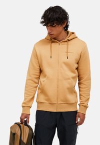 Tan zip-up hoodie med snore, kengurulommer og ribbede manchetter. Bæret over mørke bukser, holdt med en brun og beige mønstret rygsæk.