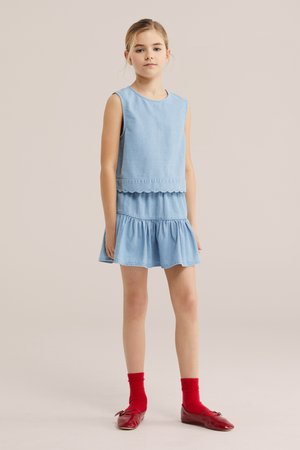 Fiatal lány áll, világoskék ujjatlan ruhát visel, hullámos szegéllyel, piros zoknival és piros lapos cipővel, egyszínű háttér előtt.