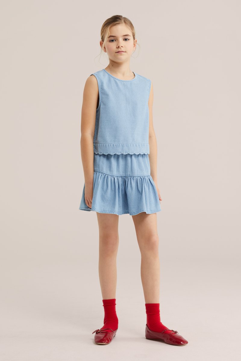 Junges Mädchen steht und trägt ein ärmelloses hellblaues Kleid mit wellenförmigem Saum, kombiniert mit roten Socken und roten flachen Schuhen vor einfachem Hintergrund.