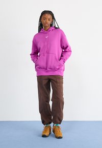 Ροζ φούτερ με μπροστινές τσέπες και κουκούλα με κορδόνι, συνδυασμένο με καφέ παντελόνι και κίτρινα αθλητικά παπού�τσια. Απλός σχεδιασμός, χαλαρή εφαρμογή.