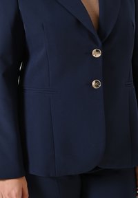 Marineblauwe op maat gemaakte blazer van gladde stof, met een enkelgeknoopte sluiting, drie metalen knopen en twee voorkruiden.