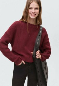 Pulover tricotat burgund cu guler rotund și manșete cu nervuri, asortat cu blugi închiși la culoare și o geantă texturată neagră cu accesorii circulare.