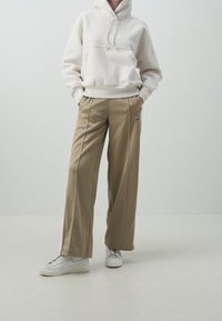 Krémová fleecová bunda s kapucí a přední kapsou, doplněná béžovými kalhotami širokého střihu se znakem Nike. Oblečeno s bílými teniskami.