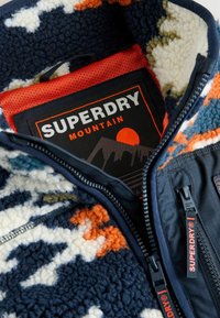 Fleecowa kurtka w wzór w granatowa, biała i pomarańczowa, czarny zamek błyskawiczny z logo Superdry oraz pomarańczowa podszewka z etykietą Superdry Mountain w środku.