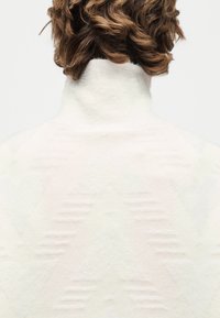 Άτομο με κοντά καστανά σγουρά μαλλιά που φοράει λευκό πουλόβερ με ψηλό λαιμό και διακριτικά υφασμένα σχέδια σε σχήμα V στην πλάτη.