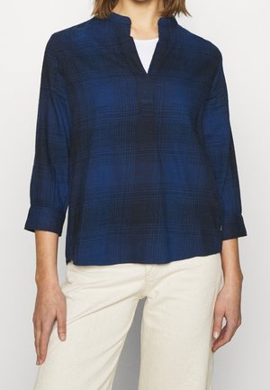 Femme portant une chemise à manches longues à carreaux bleu marine avec une encolure fendue par-dessus un haut blanc et un pantalon beige clair.