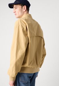 Lyse beige bomberjakke med afslappet fit, elastiske manchetter og rygdetalje, parret med blå denimjeans.