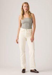 Γυναίκα που στέκεται φορώντας γκρι αμάνικη μπλούζα, λευκά τζιν ψηλόμεσα και σκουρόχρωμα καφέ παπούτσια τύπου boat, μπροστά από απλό ανοιχτόχρωμο φόντο.