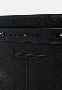 Mustro mustast nahkakott, millel on tekstuuriline pind. Omab reljeefset "TOMMY HILFIGER" logo ja väikseid trikolor aktsente. Sujuv ja stiilne disain.