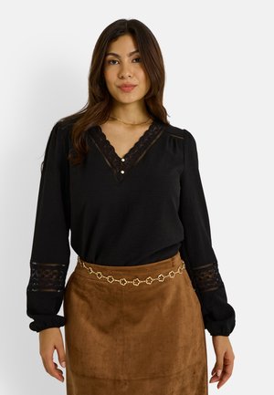 Blouse noire avec des accents en dentelle au niveau de l'encolure et des manches, dotée d'un décolleté en V et de détails de boutons. Associée à une jupe en daim marron.
