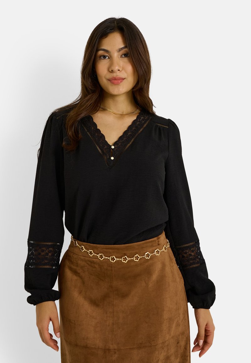 Blusa negra con detalles de encaje en el escote y las mangas, con escote en V y detalle de botones. Combinada con una falda de ante marrón.