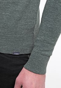 Zielony sweter z dzianiny z prążkowanym mankietem, z małą metką z napisem "1985" u dołu. Materiał wydaje się miękki i teksturowany.