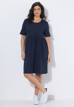 Donna con capelli ricci scuri che indossa un vestito blu navy fino al ginocchio e scarpe da ginnastica bianche, in piedi con le gambe incrociate davanti a uno sfondo bianco.