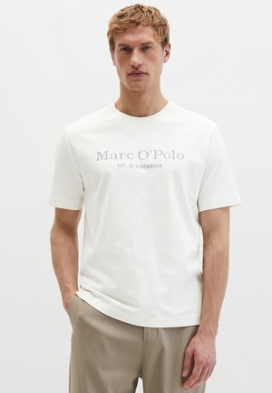 Blonde man met een wit Marc O'Polo T-shirt met de tekst "EST. IN STOCKHOLM" en beige broek, staand tegen een effen achtergrond.