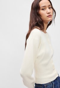 Κρέμα ζακέτα με ριμπ τιράντες και μακριά μανίκια, με κουμπιά μπροστά και μαλακή πλέξη. Συνδυάζεται με τζιν παντελόνι.