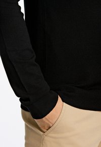 Maglione nero aderente a manica lunga con polsini a coste, abbinato a pantaloni beige chiaro. Il maglione ha una texture liscia e un design semplice.