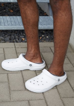 White slip-on clogs with ventilation holes, textured soles, and a gray interior lining, placed on a paved surface.