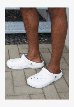 Valkoiset liukuva kenkäklossit, joissa on ilmanvaihtoaukkoja, teksturoidut pohjat ja harmaa sisus, asetettuna päällystetylle pinnalle.