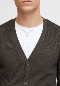 Mørk olivengrønn cardigan med ribbestrikkede skuldre, V-halsdesign og to knapper. Brukt over en hvit T-skjorte, med en sølvkjede med anheng.