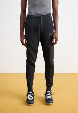 Personne portant un jogging noir Nike avec une taille à cordon et des baskets noires et blanches, debout sur un sol jaune contre un mur blanc.