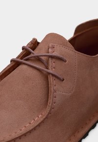 Κοντινό πλάνο ενός ροζ παπουτσιού από σουέντ με ραμμένες λεπτομέρειες και δεμένα στρογγυλά κορδόνια σε απλό φωτεινό φόντο.