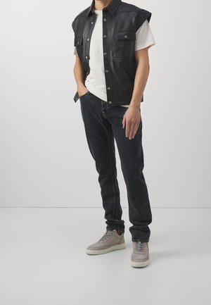 Henkilö, joka pukeutuu mustaan hihattomaan nahkaliiviin valkoisen t-paidan päällä, tummiin farkkuihin ja vaaleanharmaisiin lenkkareihin, seisten käsi taskussa.