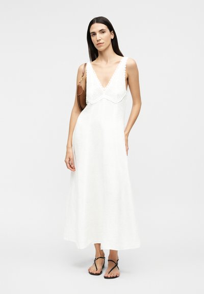 Nő ujjatlan fehér maxi ruhában, amely horgolt részletekkel díszített, barna válltáskát visel és fekete szandált hord.
