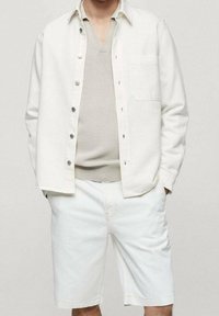 Camicia bianca a maniche lunghe sopra un maglione beige a scollo a V, abbinata a pantaloncini bianchi in denim. La camicia presenta una singola tasca sul petto e bottoni argentati.