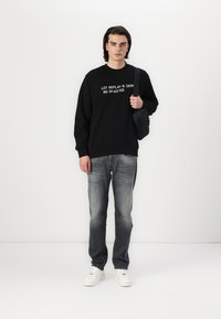Sudadera negra con texto gráfico blanco, combinada con jeans grises y zapatillas blancas, sosteniendo un bolso negro, de pie sobre un fondo simple.