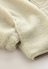 Tkanina z kremowego polaru o miękkiej, teksturowanej powierzchni. Posiada gładkie, kontrastowe beżowe wykończenie na dole i otworach rękawów.