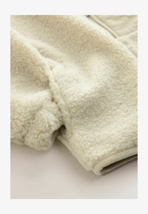 Fleecani materijal krem boje s mekom, teksturiranom površinom. Ima glatki, kontrastni bež obrub duž dna i otvorâ rukava.