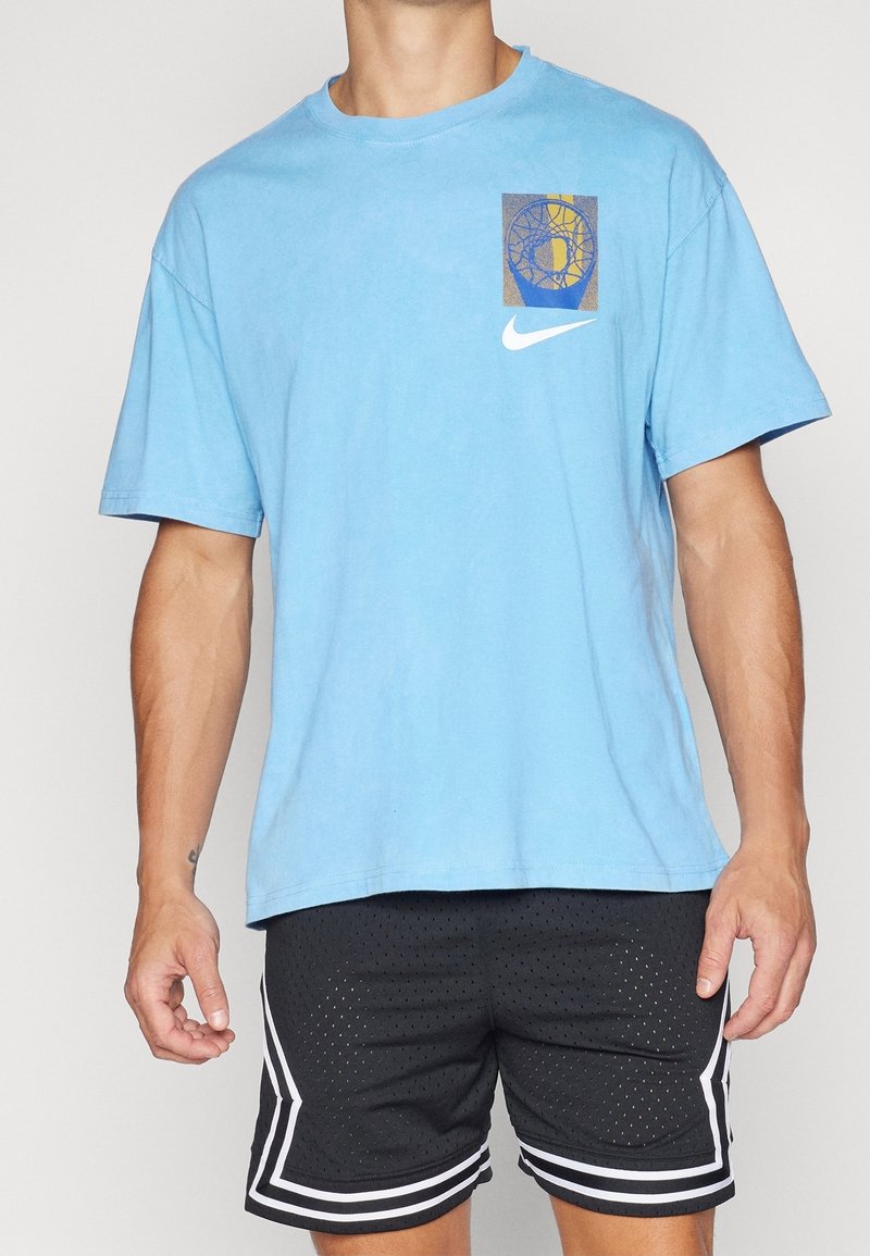 Homme portant un t-shirt Nike bleu clair avec un graphisme de panier de basket et un short de sport noir perforé avec des bandes blanches.