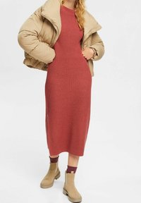 Veste matelassée beige, robe en maille côtelée couleur rouille, et bottines couleur beige avec semelles en caoutchouc. La robe présente une coupe ajustée et une longueur midi.